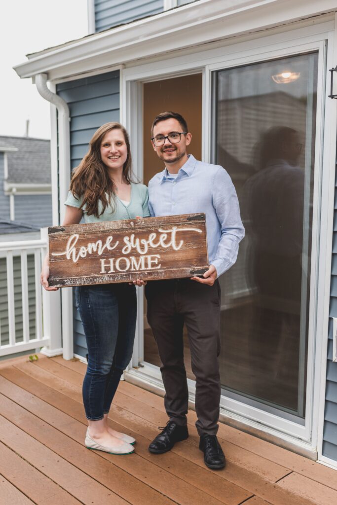 Smiling couple holding 'Home Sweet Home' sign in front of their new house.